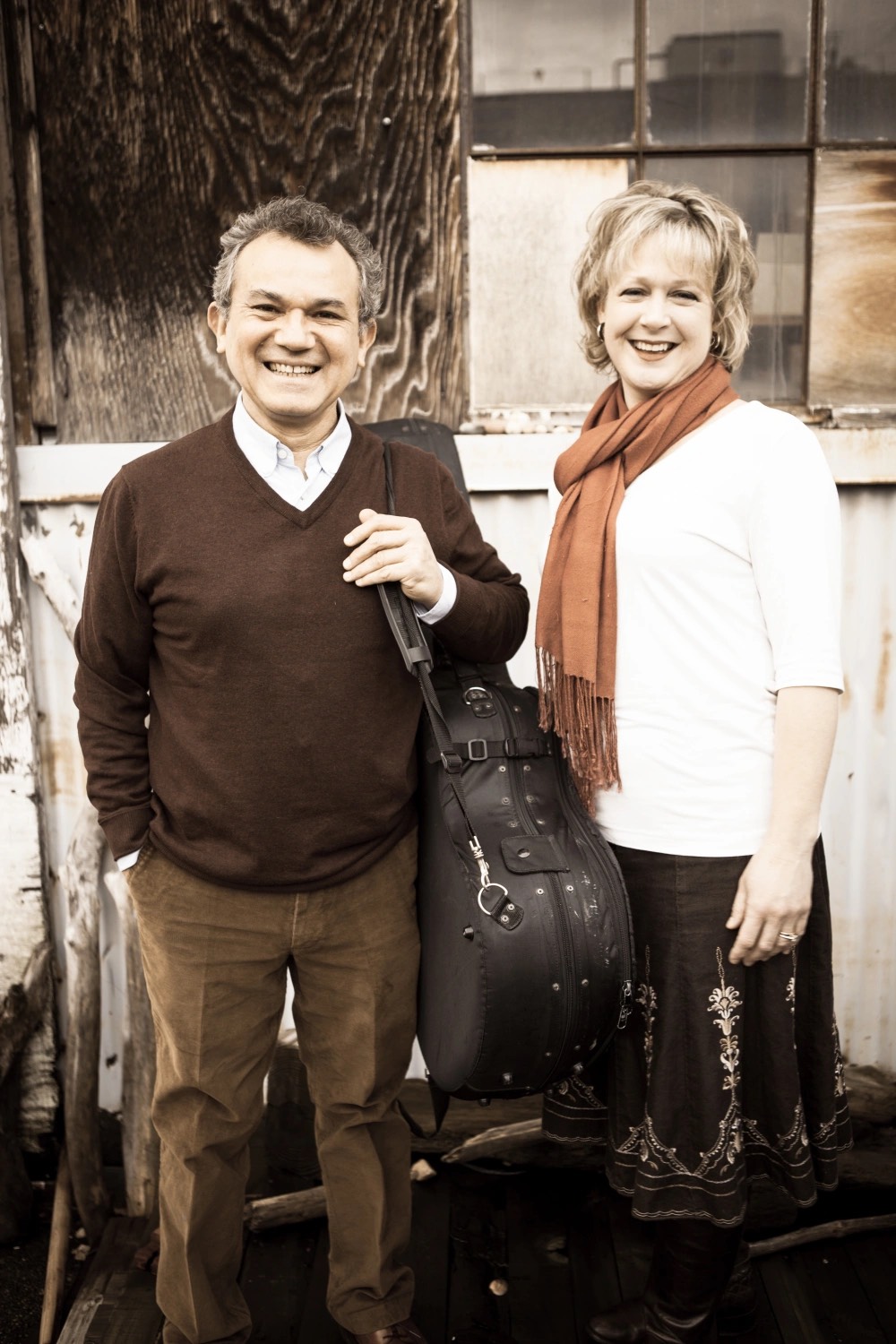 Chérie L. Hughes and guitarist Roberto Limón of The Pacific Duo, smiling in front of a weathered wall, Roberto holding his guitar case
