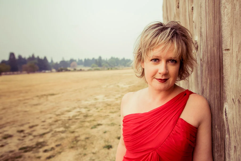Chérie L. Hughes in a red one-shoulder gown, leaning against a weathered barn wall beside an open field